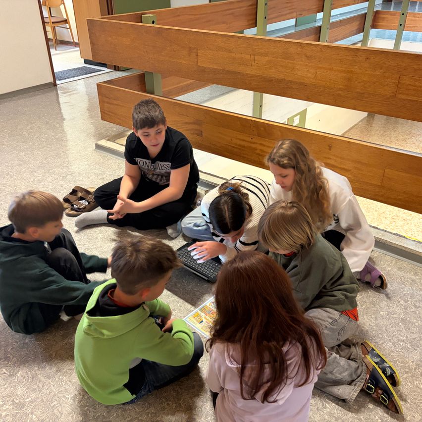 Eine Gruppe von Schülern sitzt auf dem Boden und benutzt einen Laptop. Einige schauen in ein Buch, andere auf den Laptop.