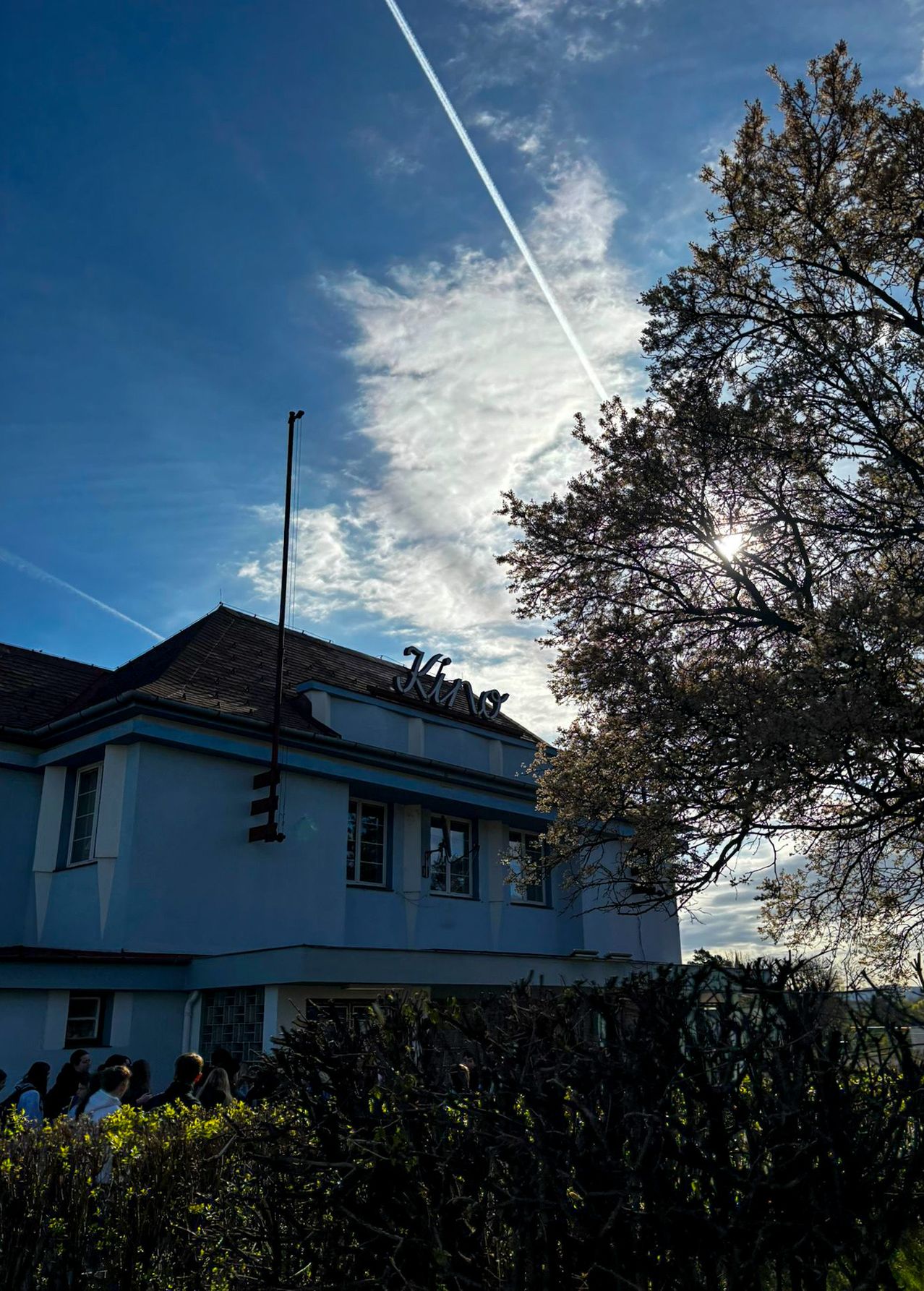 Ein weißes Gebäude mit dem Wort Kinos oben ist von einem Baum und Büschen umgeben. Menschen versammeln sich vor dem Gebäude unter einem teilweise bewölkten Himmel.