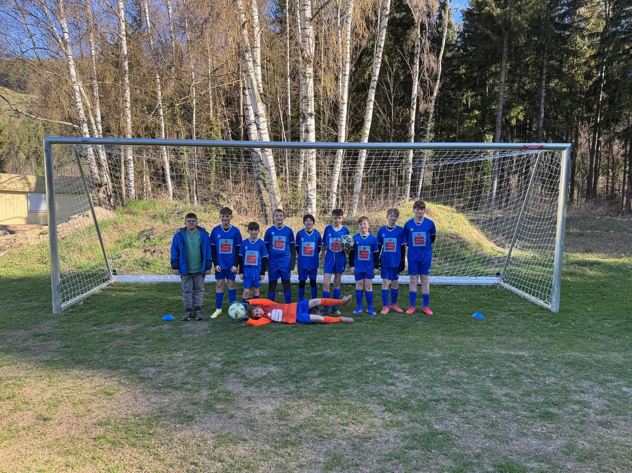 Eine Gruppe junger Fußballspieler in blauen Uniformen posiert für ein Foto auf einem Feld, wobei ein Spieler in der Nähe eines Fußballs auf dem Boden liegt.