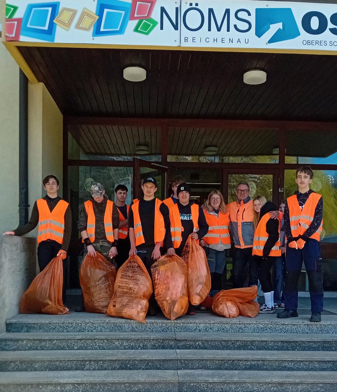 Eine Gruppe von Freiwilligen in Sicherheitswesten steht vor einem Gebäude mit mehreren Mülltüten. Sie haben sich für eine Aufräumaktion versammelt.