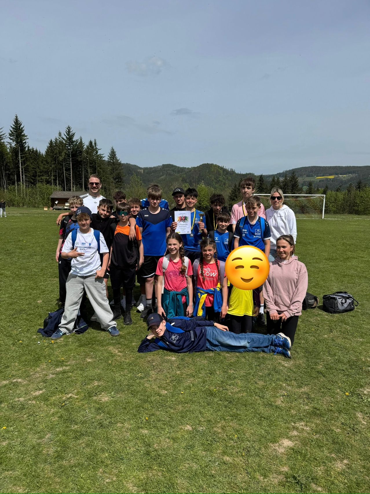 Eine Gruppe von Kindern und Erwachsenen posiert für ein Foto auf einem Rasenfeld. Einige halten Trophäen, während eine Person auf dem Boden liegt. Bäume und ein Berg sind im Hintergrund.