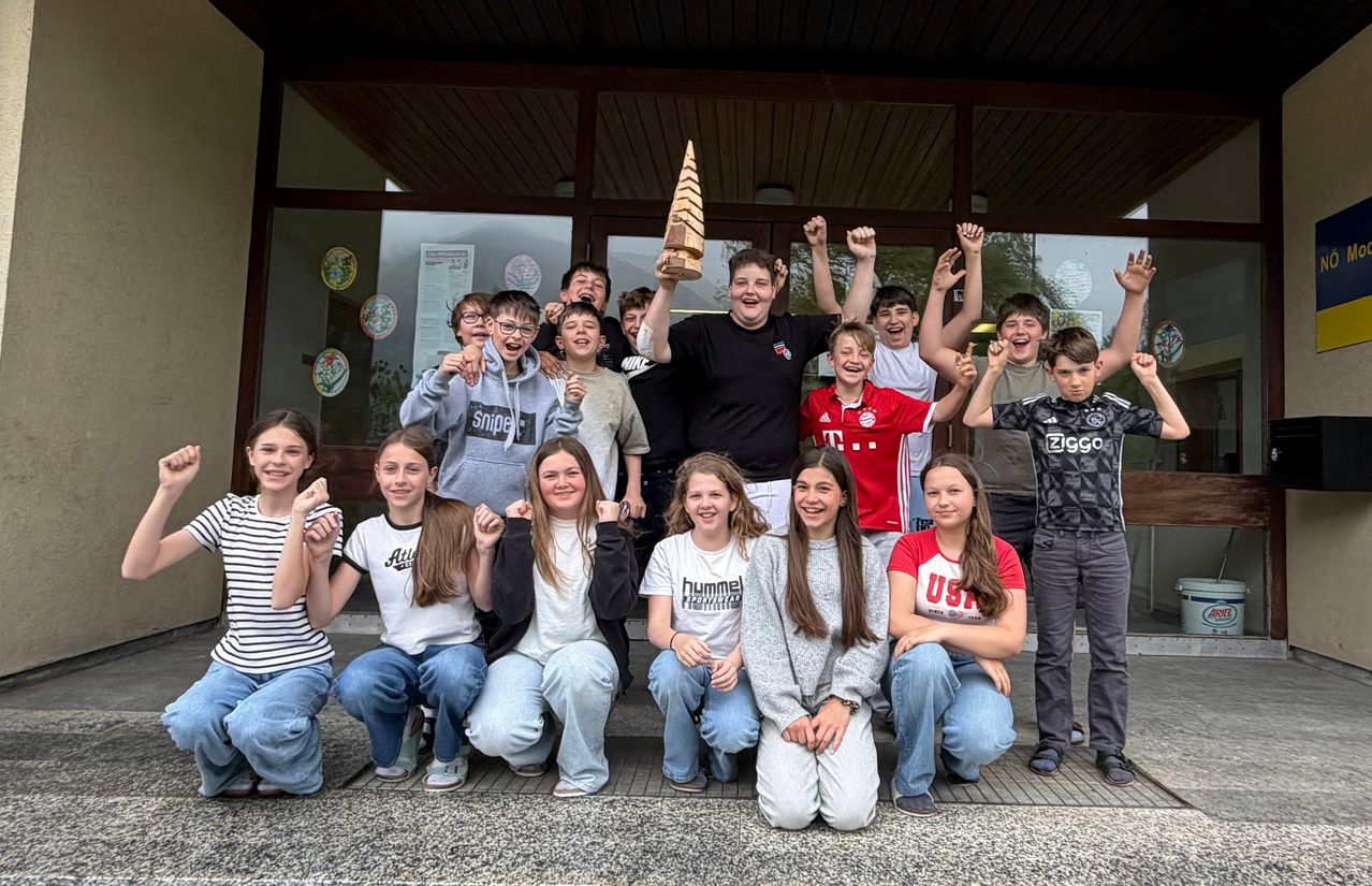 Eine Gruppe von Teenagern posiert für ein Foto vor einem Gebäude. Einige sitzen, andere stehen. Ein Junge in der Mitte hält einen Holzkegel. Hinter ihnen sind Glasfenster mit einigen runden Aufklebern.