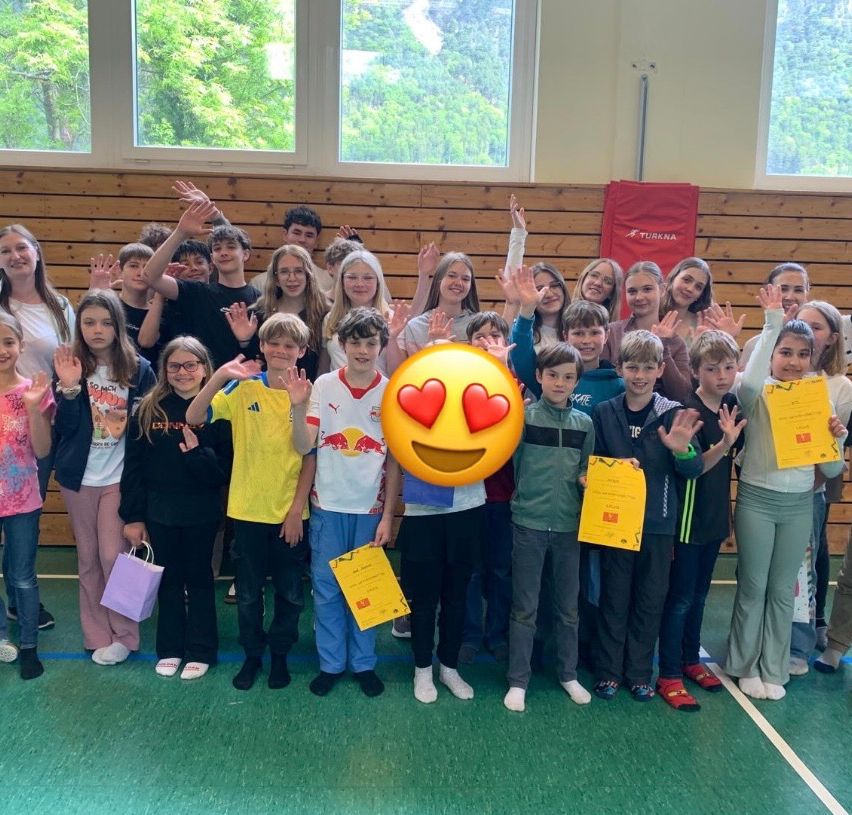 Gruppenfoto von Schülern in einer Turnhalle, einige halten Urkunden, lächeln und machen Handzeichen, mit einem Herz-Emoji in der Mitte.