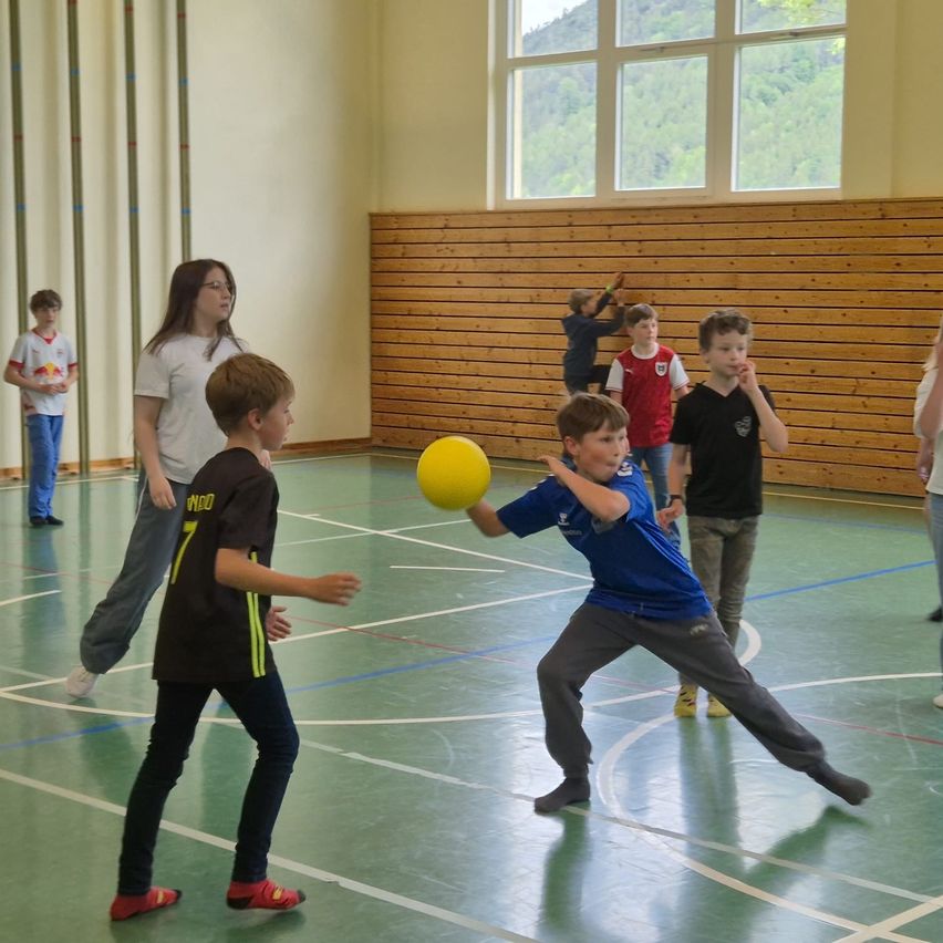 Mehrere Kinder spielen mit einem gelben Ball in einer Turnhalle. Zwei Jungen spielen aktiv, während andere zusehen, und eine Frau steht hinter ihnen. Die Turnhalle hat eine Holzwand und Glasfenster.