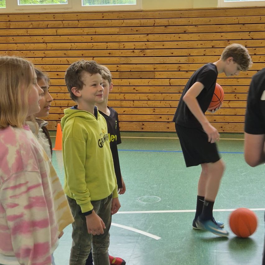 Several children watch a boy in a black shirt dribble a basketball indoors on a green court. Behind them, a wooden wall and a traffic cone.