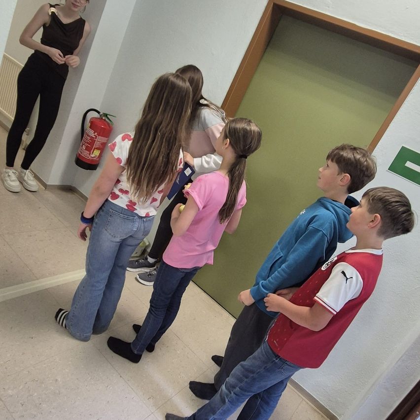 Five children stand near a green door, looking at something. A woman stands behind them. A fire extinguisher is mounted on the wall.