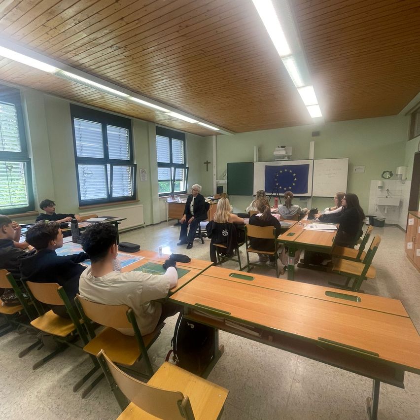 Ein Klassenzimmer mit Schülern und einem Lehrer, die in einem Kreis sitzen. Eine europäische Flagge ist an der Wand hinter dem Lehrer angebracht.