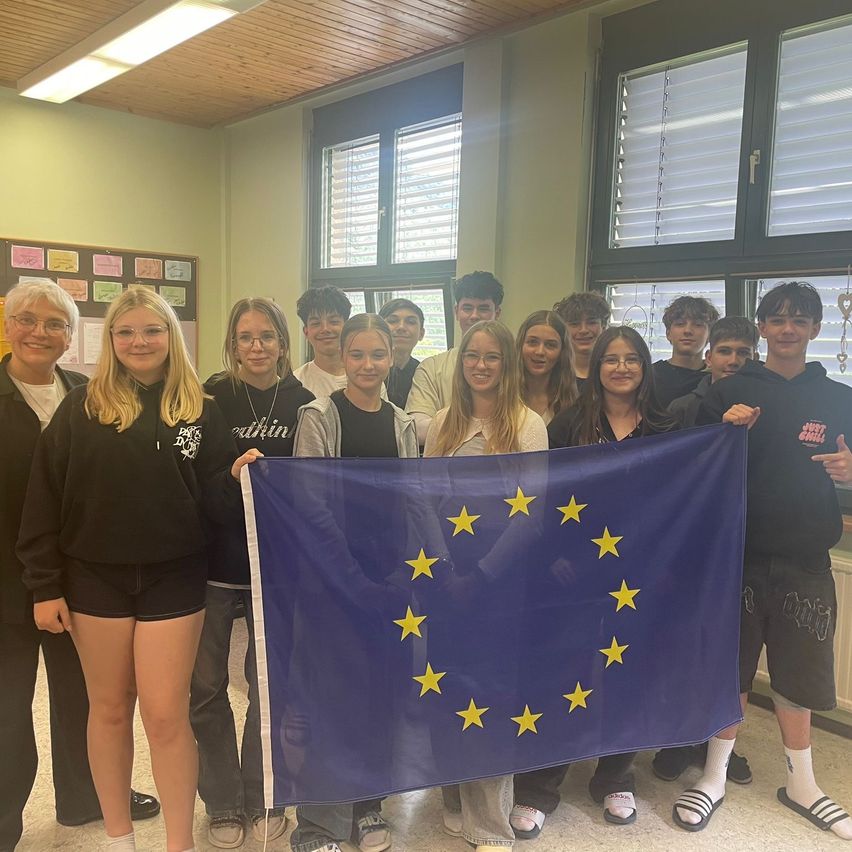 Eine Gruppe junger Menschen steht in einem Klassenzimmer und hält eine Flagge der Europäischen Union. Sie alle lächeln und posieren für ein Foto.