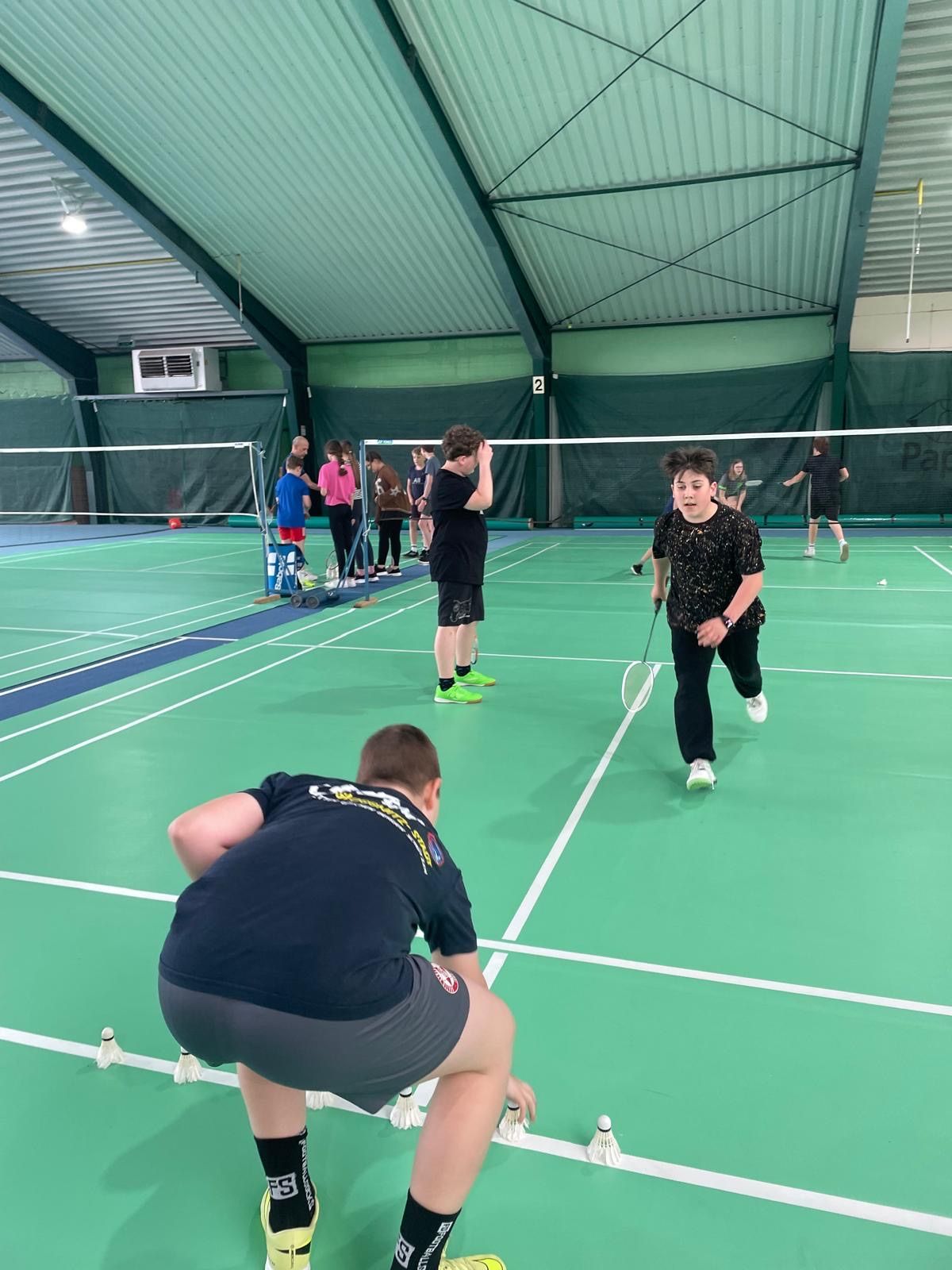 Eine Gruppe junger Leute spielt Badminton auf einem Hallenplatz. Einer beugt sich nach vorne, um einen Federball aufzuheben, während ein anderer sich auf den Aufschlag vorbereitet.