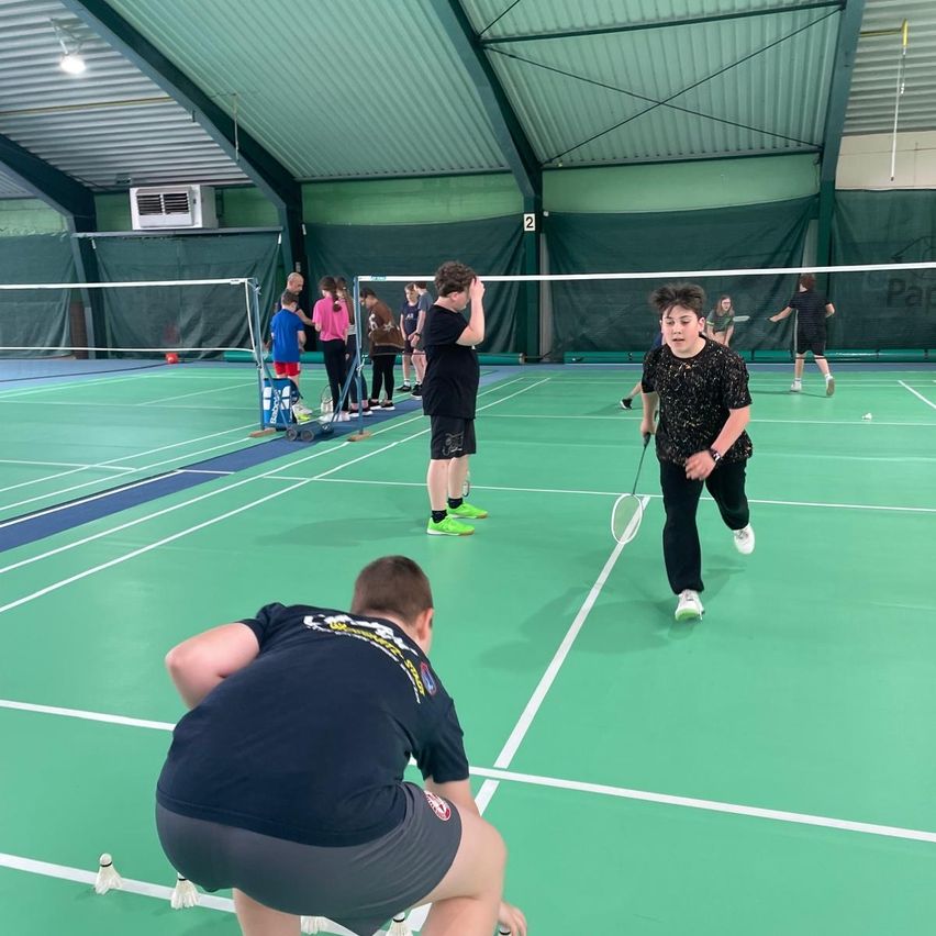 Eine Gruppe junger Leute spielt Badminton auf einem Hallenplatz. Einer beugt sich nach vorne, um einen Federball aufzuheben, während ein anderer sich auf den Aufschlag vorbereitet.