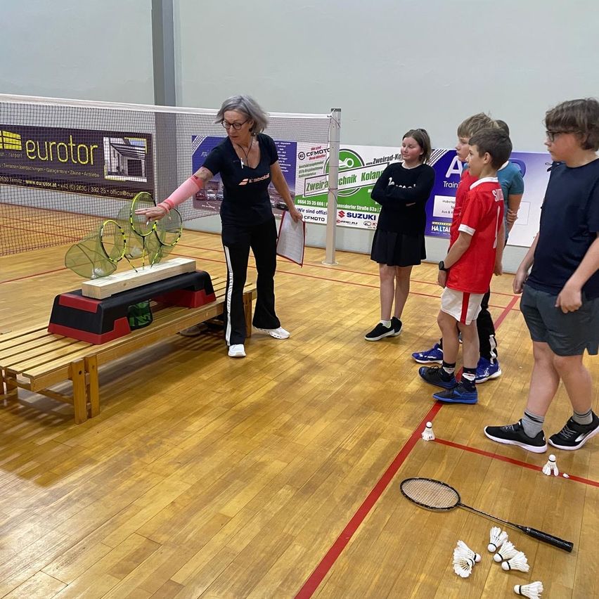 Eine Trainerin demonstriert eine Badminton-Einrichtung mit Schlägern auf einer Bank. Eine Gruppe von Kindern und einer Frau steht in der Nähe und beobachtet. Der Platz ist mit roten Linien markiert.