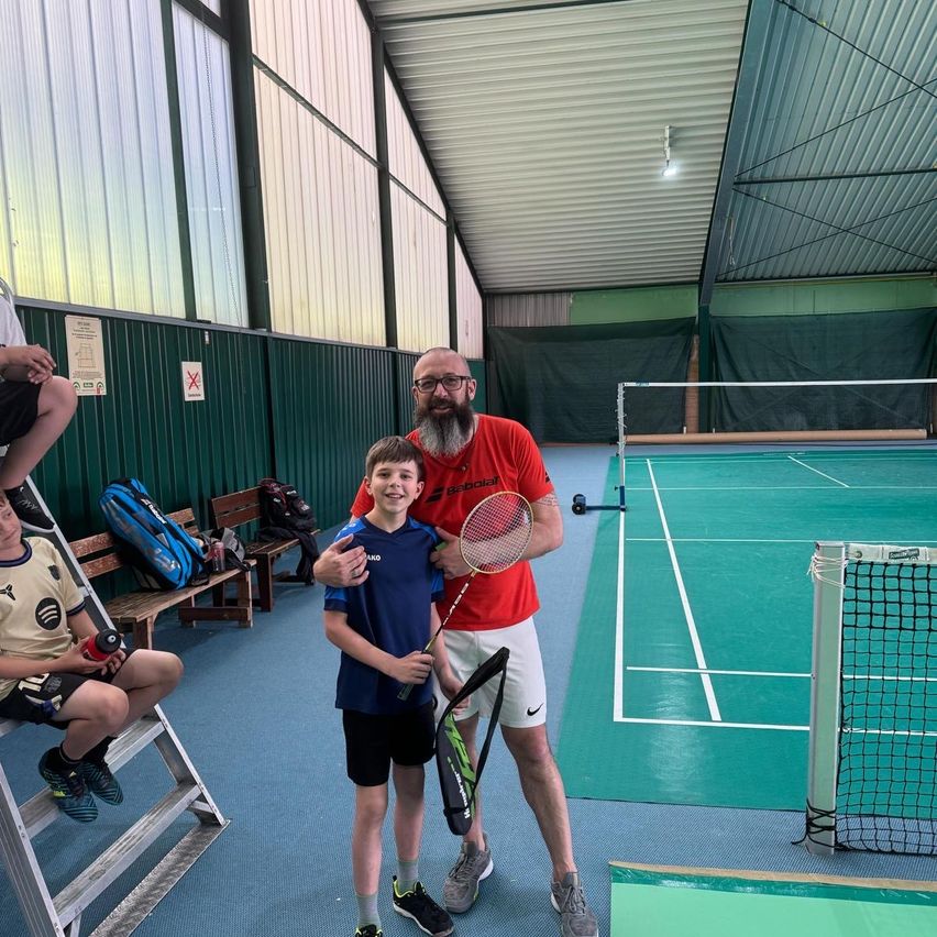 Ein Mann und ein Junge stehen zusammen auf einem Badmintonplatz und halten Schläger. Der Junge trägt ein blaues Shirt und schwarze Shorts. Ein anderer Junge sitzt auf einer Leiter in der Nähe.