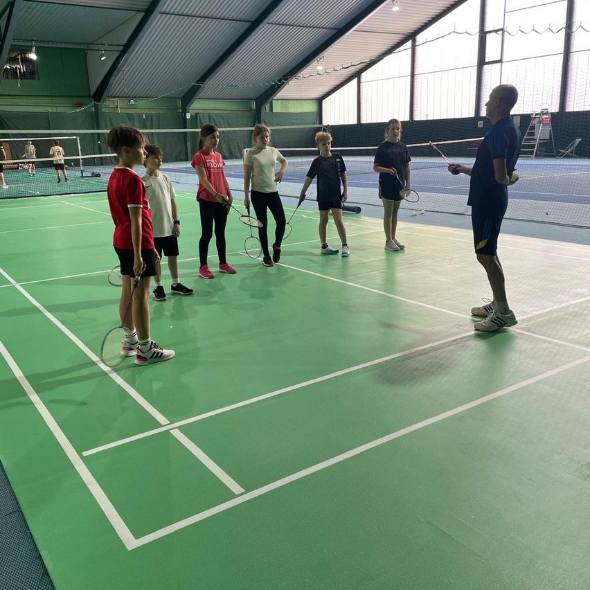 Eine Gruppe junger Kinder und ein Mann stehen auf einem Badminton-Court, halten Schläger und haben zwei Netze aufgestellt. Der Platz ist grün mit weißen Linien.