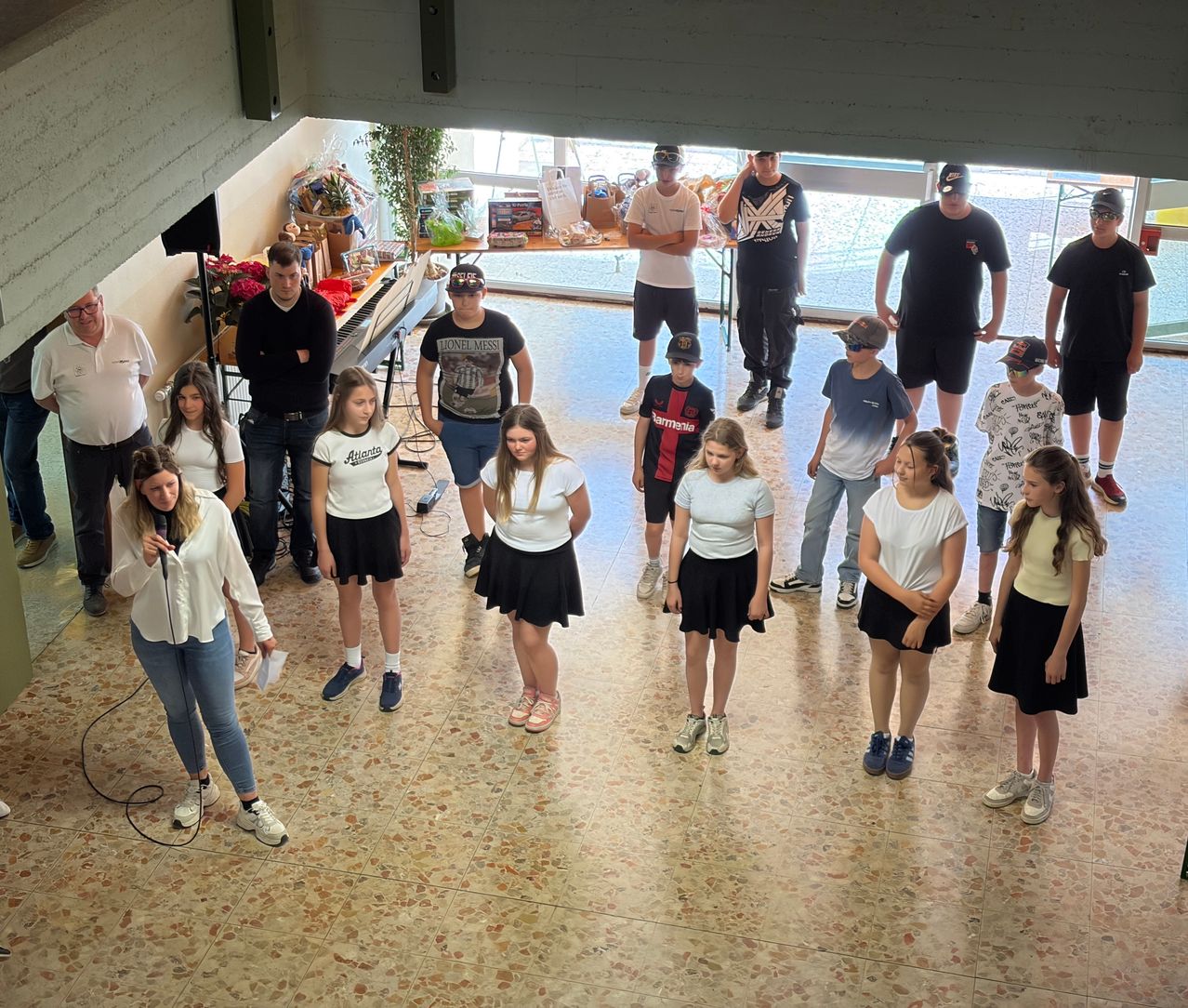 Eine Gruppe von Menschen versammelt sich drinnen; einige tragen Hüte, Turnschuhe und Brillen. Eine Frau spricht in ein Mikrofon, während andere vor ihr stehen. Ein Tisch mit Gegenständen steht hinter ihnen.