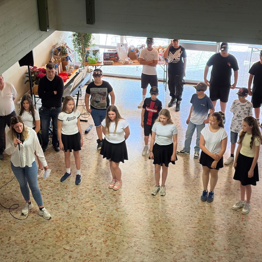Eine Gruppe von Menschen versammelt sich drinnen; einige tragen Hüte, Turnschuhe und Brillen. Eine Frau spricht in ein Mikrofon, während andere vor ihr stehen. Ein Tisch mit Gegenständen steht hinter ihnen.