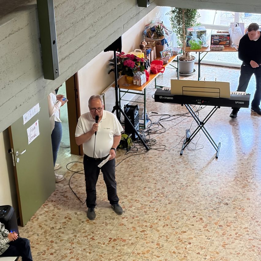 Ein älterer Mann spricht in ein Mikrofon. Hinter ihm steht ein Klavier. Ein weiterer Mann steht bei einem Tisch mit Blumen. Eine Frau steht in der Tür.