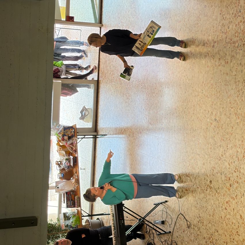 Eine Frau mit Mikrofon und Klavier singt in einem Raum mit gefliesten Böden und einer Glaswand. Eine weitere Frau hält ein Buch und steht hinter ihr.