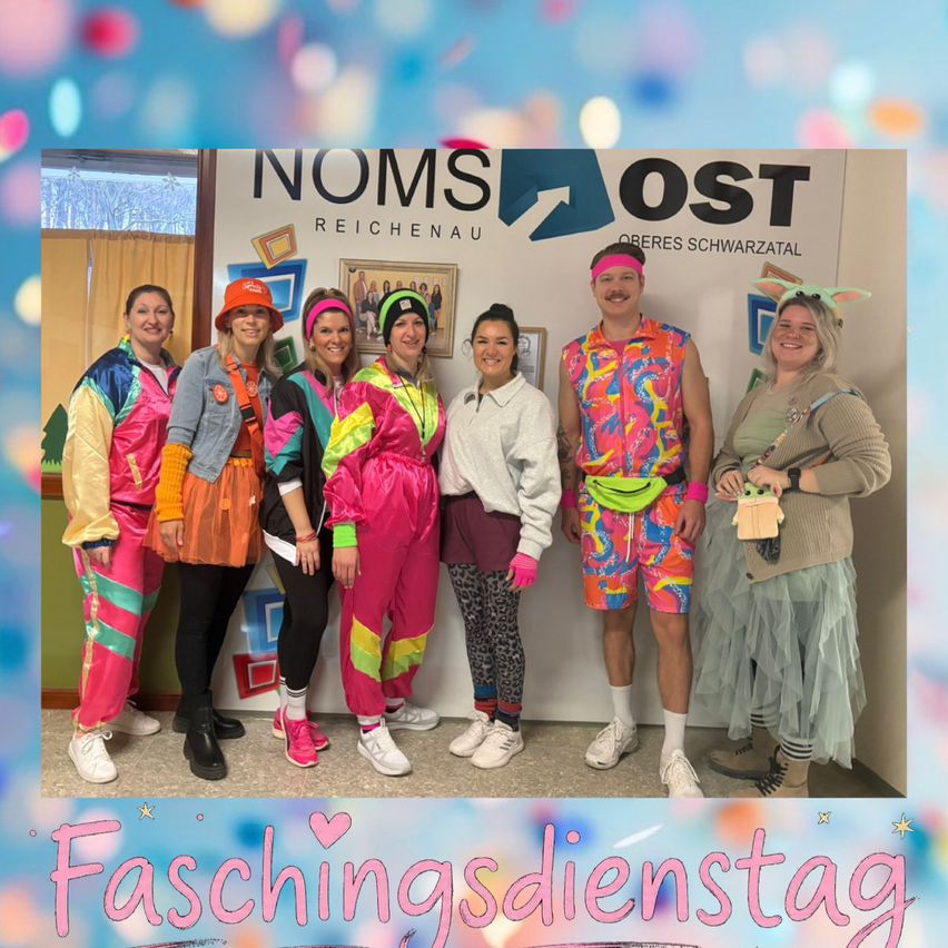 Eine Gruppe von Menschen in leuchtenden, farbenfrohen Kostümen posiert für ein Foto vor einem Schild, das 'Faschingsdienstag' steht. Sie lächeln und scheinen ein Ereignis zu feiern.