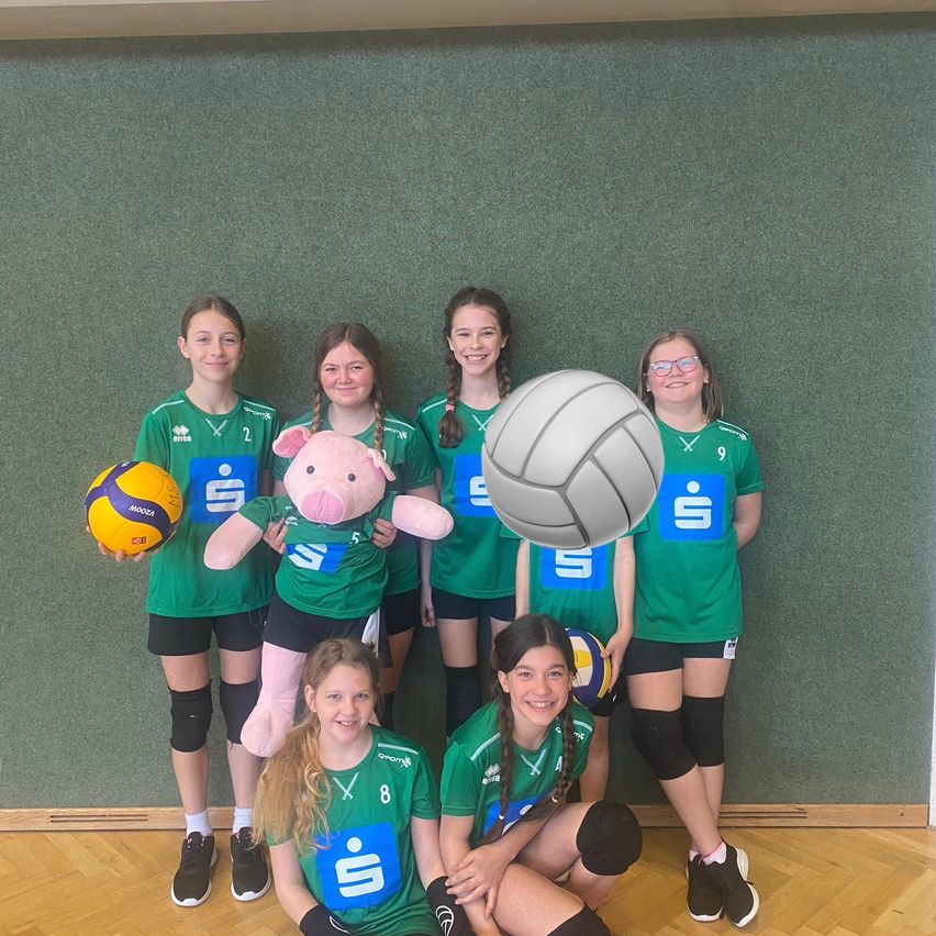 Eine Gruppe von Mädchen in grünen Trikots steht zusammen, eine hält einen Ball und ein rosa Schweinchen ist in der Mitte. Sie lächeln und posieren für ein Foto.