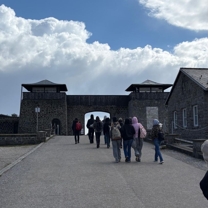 Eine Gruppe von Menschen geht auf den Eingang eines historischen Steingebäudes zu. Der Himmel ist blau mit verstreuten Wolken.