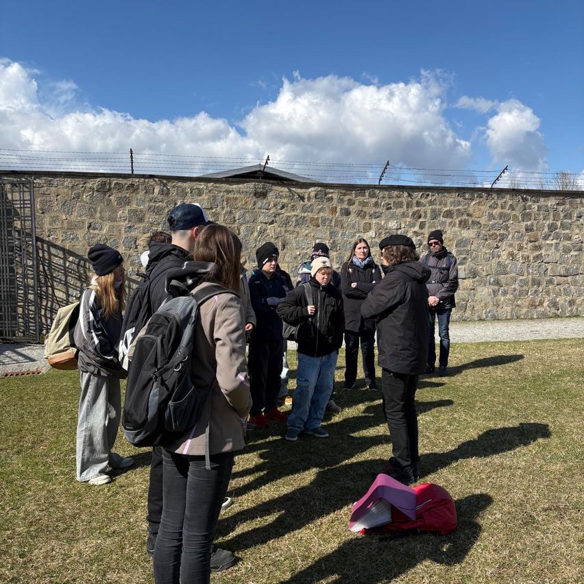 Eine Gruppe von Menschen steht vor einer Steinmauer, einige tragen Mützen und haben Rucksäcke dabei, und lauschen einer Frau mit Mikrofon.
