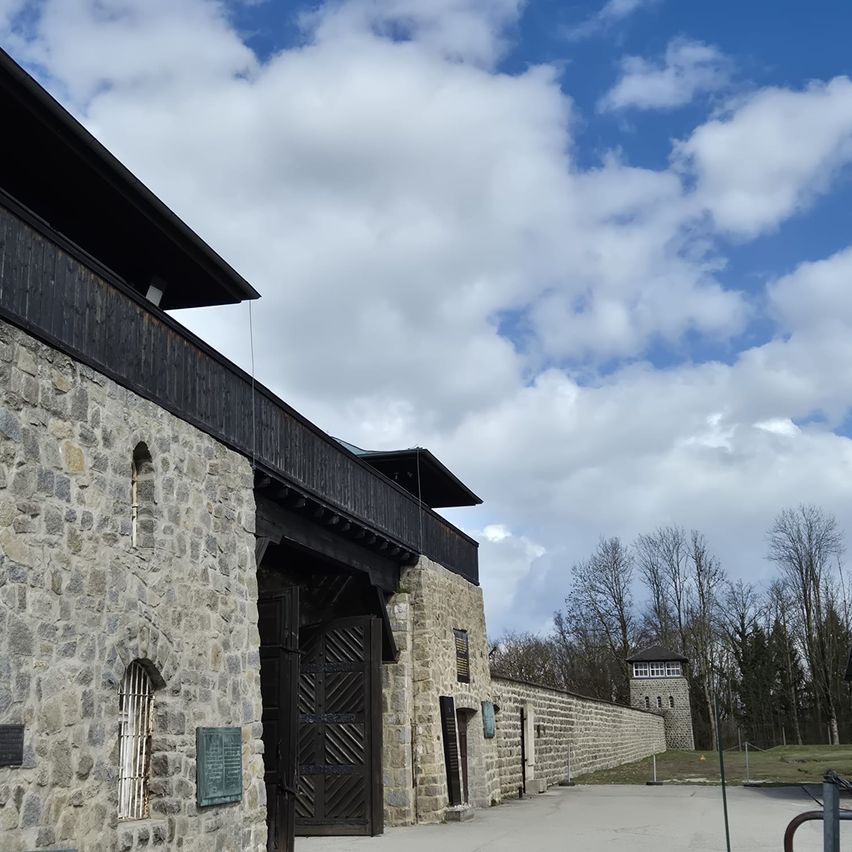 Ein Steingebäude mit einer großen Holztür unter einem blauen Himmel mit Wolken. Im Hintergrund befindet sich ein Zaun und Bäume.