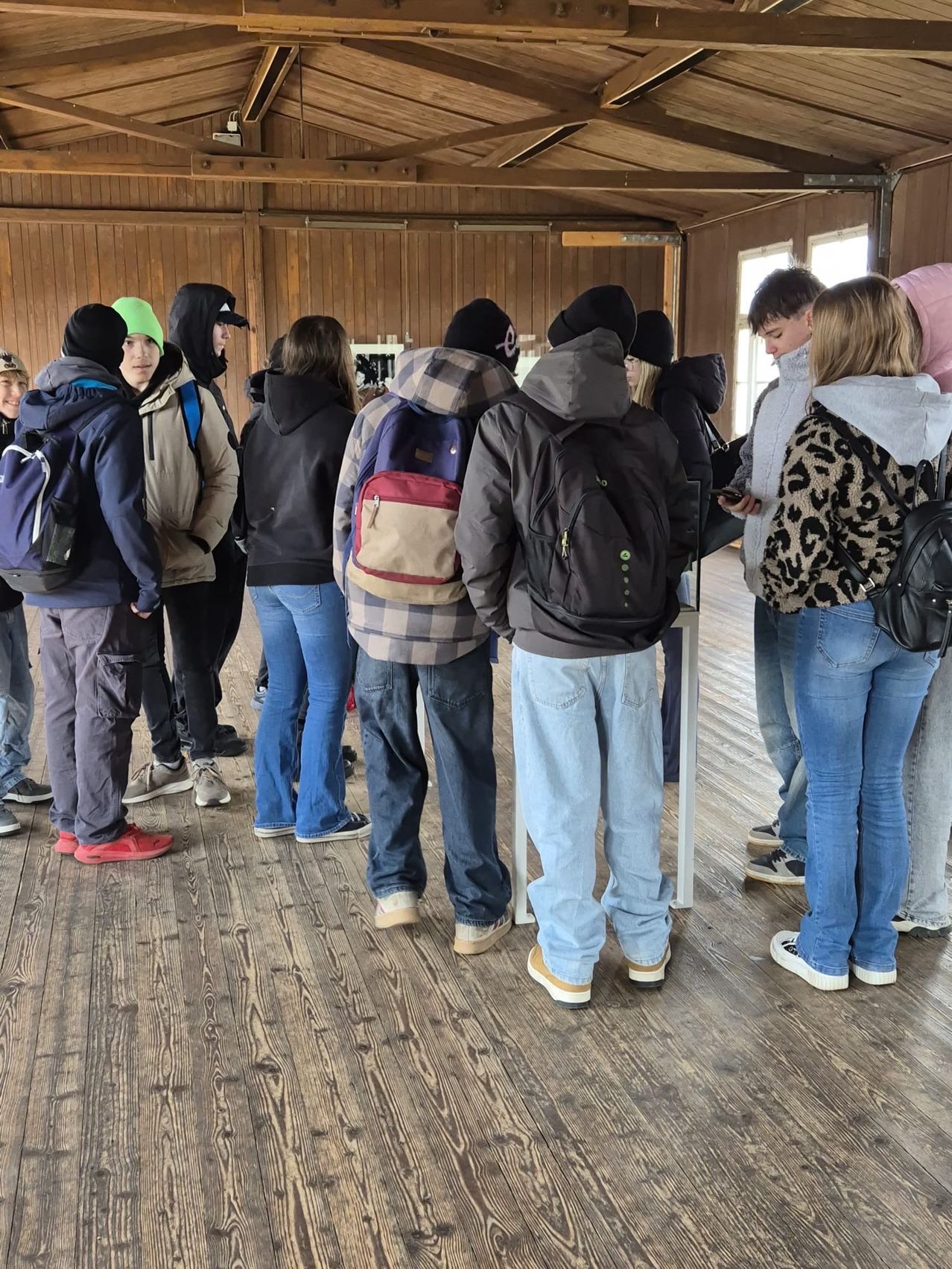 Eine Gruppe von Menschen in Winterkleidung steht in einer Schlange in einem hölzernen Gebäude mit gefliestem Boden. Einige tragen Rucksäcke und benutzen ihre Handys.