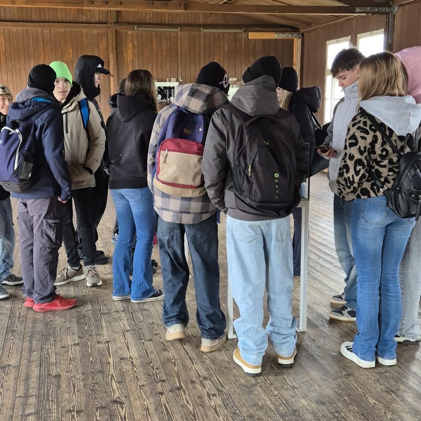 Eine Gruppe von Menschen in Winterkleidung steht in einer Schlange in einem hölzernen Gebäude mit gefliestem Boden. Einige tragen Rucksäcke und benutzen ihre Handys.
