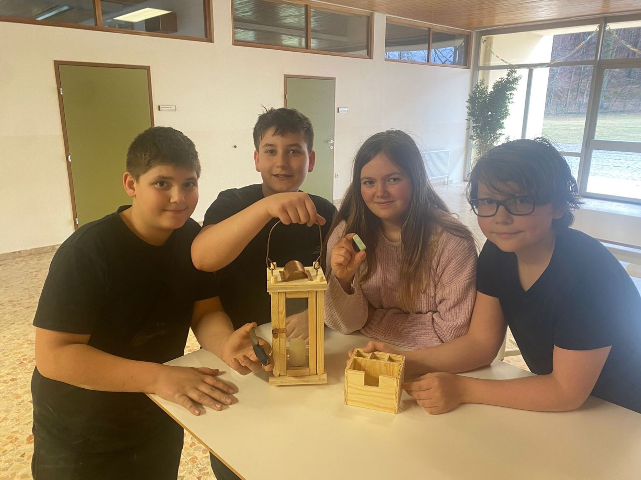 Vier Schüler sitzen lächelnd um einen Tisch herum. Sie halten eine kleine Laterne und eine Holzkiste. Der Raum hat weiße Wände und Glasfenster, mit einer Topfpflanze im Hintergrund.