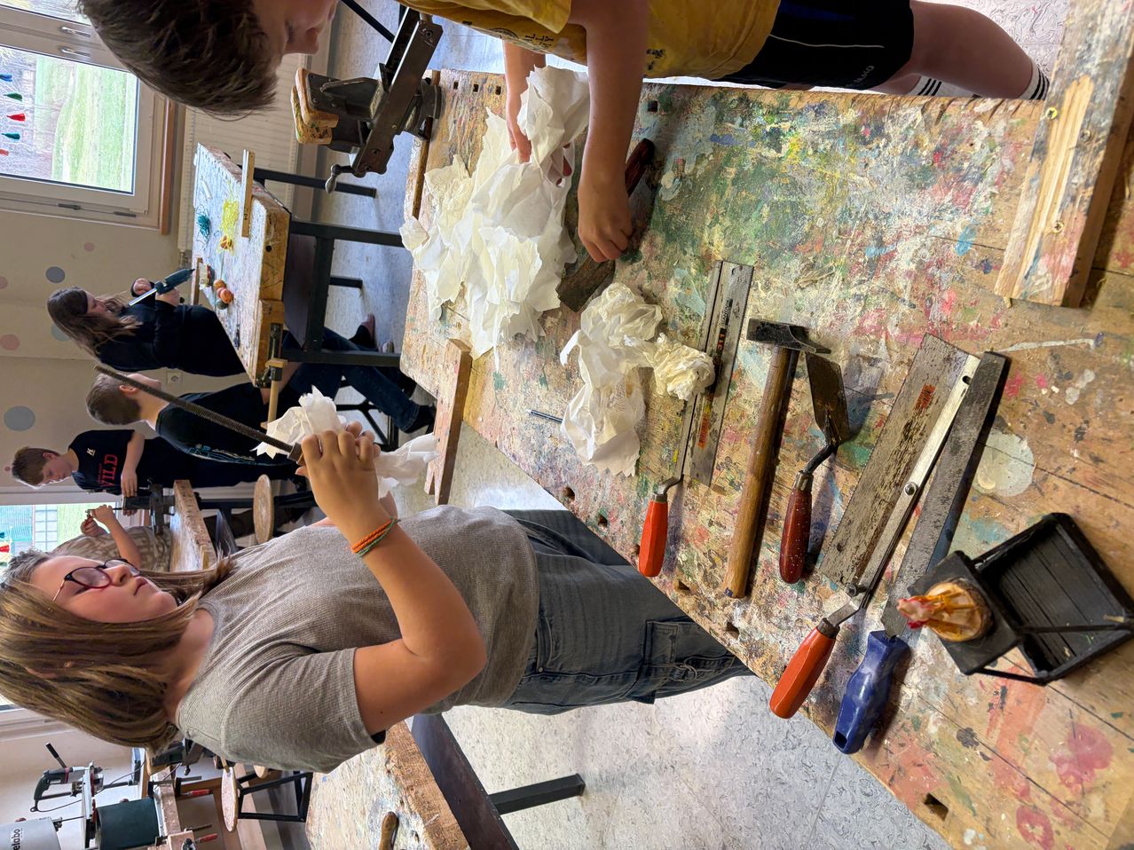 Mehrere Kinder in einer Werkstatt, die Werkzeuge und Materialien auf einem unordentlichen Tisch verwenden, ein Kind mit einem Pinsel.