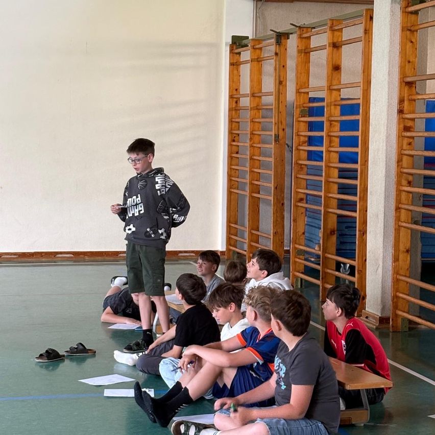 Ein Junge steht in einer Turnhalle und spricht eine Gruppe sitzender Jungen an. Die Jungen hören aufmerksam zu. Einige Schuhe und Papiere liegen auf dem Boden.