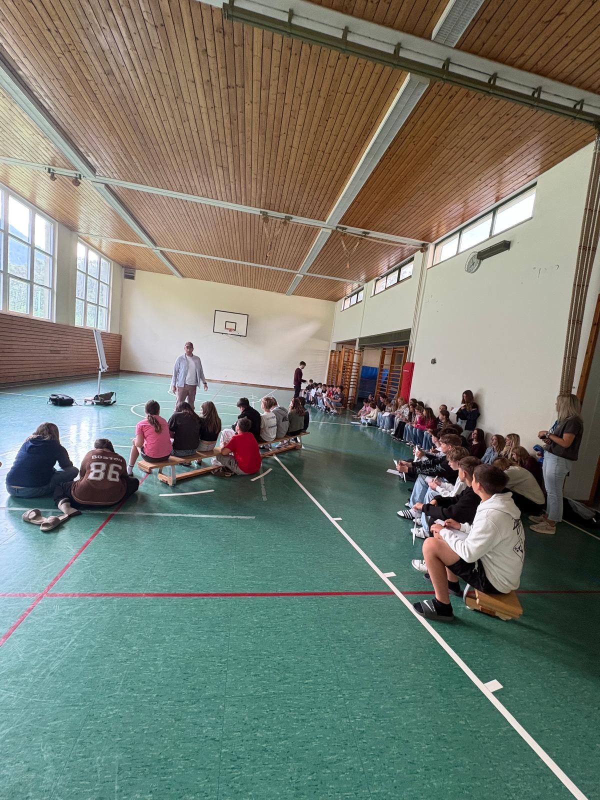 In einer Sporthalle sitzen eine Gruppe von Schülern und einem Lehrer auf Bänken, mit einem Mann in der Mitte. Ein Basketballkorb ist an der Wand montiert. Einige Schüler stehen in der Nähe der Wand.