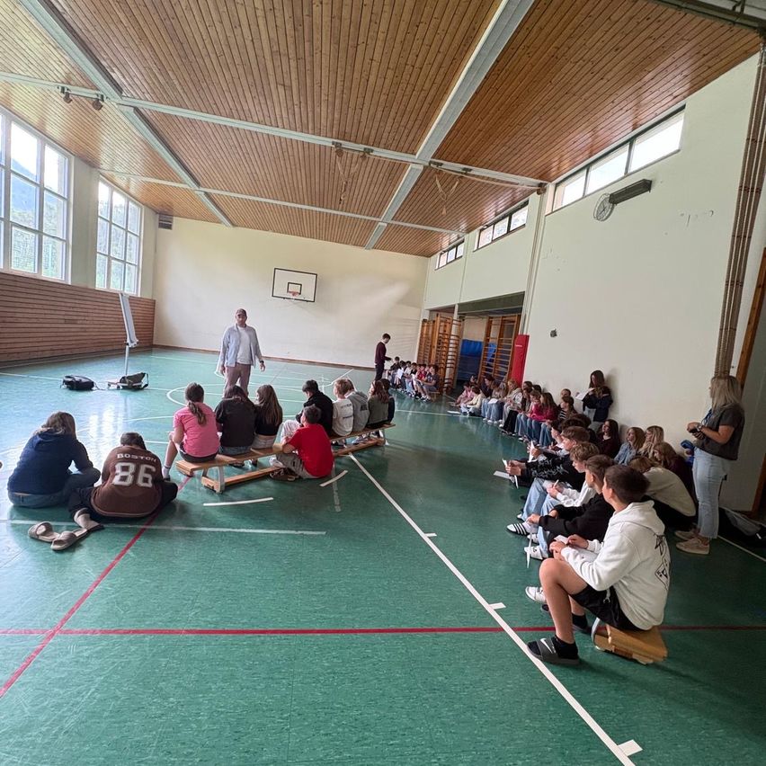In einer Sporthalle sitzen eine Gruppe von Schülern und einem Lehrer auf Bänken, mit einem Mann in der Mitte. Ein Basketballkorb ist an der Wand montiert. Einige Schüler stehen in der Nähe der Wand.