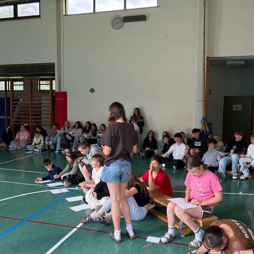 In einer Turnhalle steht eine Frau vor einem sitzenden Publikum und hält ein Papier in der Hand. Das Publikum ist engagiert, schreibt auf Papieren und hört aufmerksam zu. Der Raum hat einen grünen Boden, weiße Wände und ein rotes Banner.