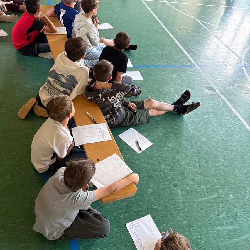 Mehrere Jungen sitzen auf einer Bank in einer Turnhalle und schreiben auf Papier. Sie sind lässig gekleidet, und einige tragen Sandalen. Der Hallenboden hat Markierungen.