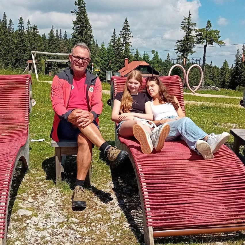 Ein älterer Mann und zwei Mädchen sitzen auf roten Liegestühlen auf einer Wiese, umgeben von Kiefernbäumen und einem bewölkten Himmel.