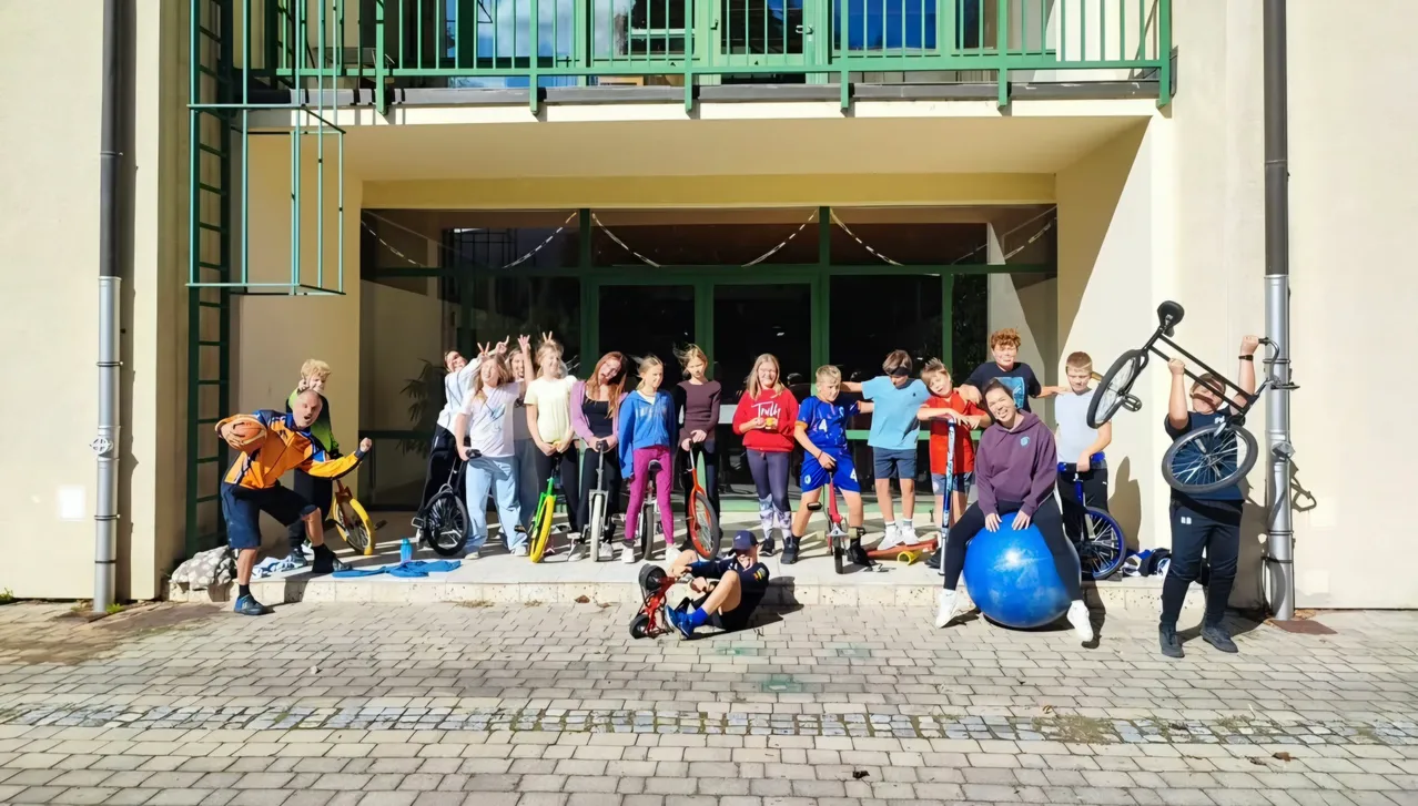 Eine Gruppe von Menschen posiert für ein Foto vor einem Gebäude mit einem gepflasterten Boden. Einige von ihnen halten Fahrräder und Bälle, während andere auf dem Boden sitzen.