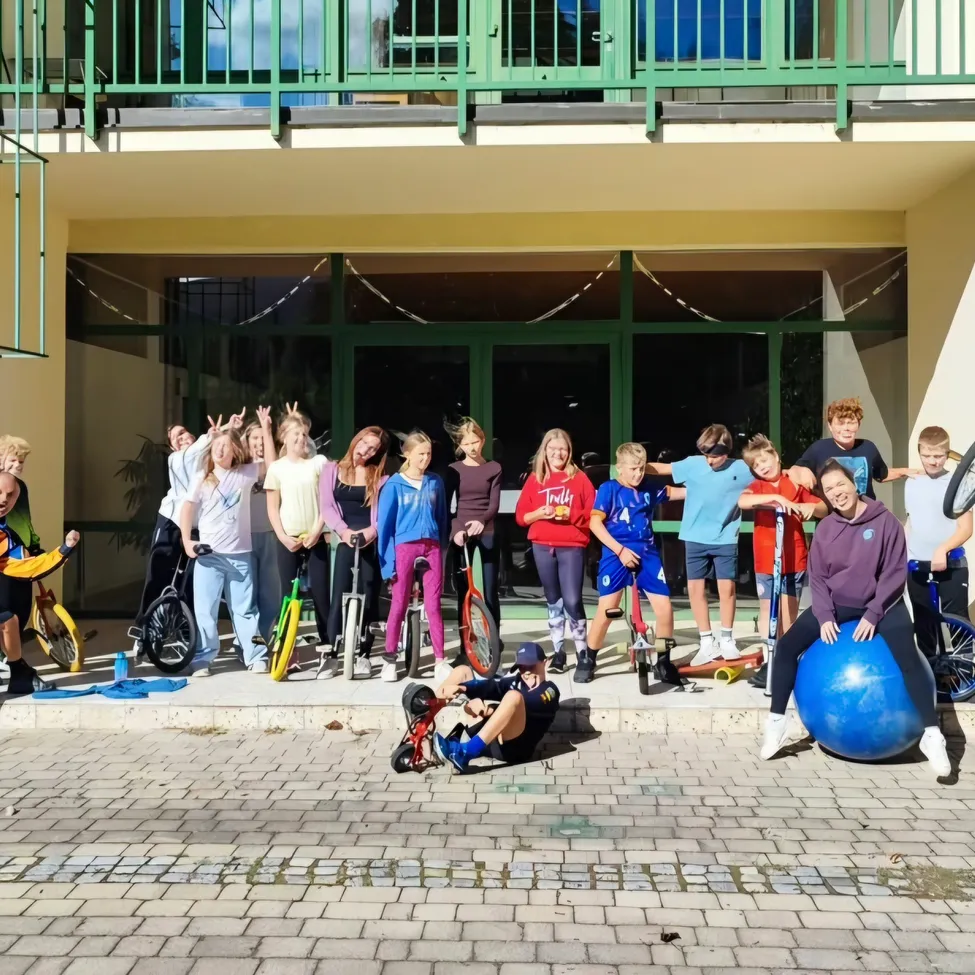 Eine Gruppe von Menschen posiert für ein Foto vor einem Gebäude mit einem gepflasterten Boden. Einige von ihnen halten Fahrräder und Bälle, während andere auf dem Boden sitzen.