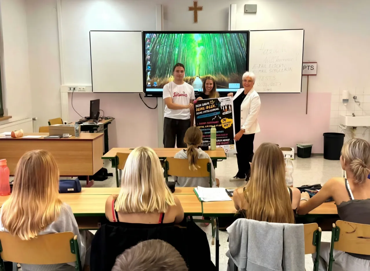 Eine Gruppe von Menschen ist in einem Klassenzimmer. Zwei Frauen und ein Mann stehen vor einer großen Leinwand. Der Mann hält ein Plakat.