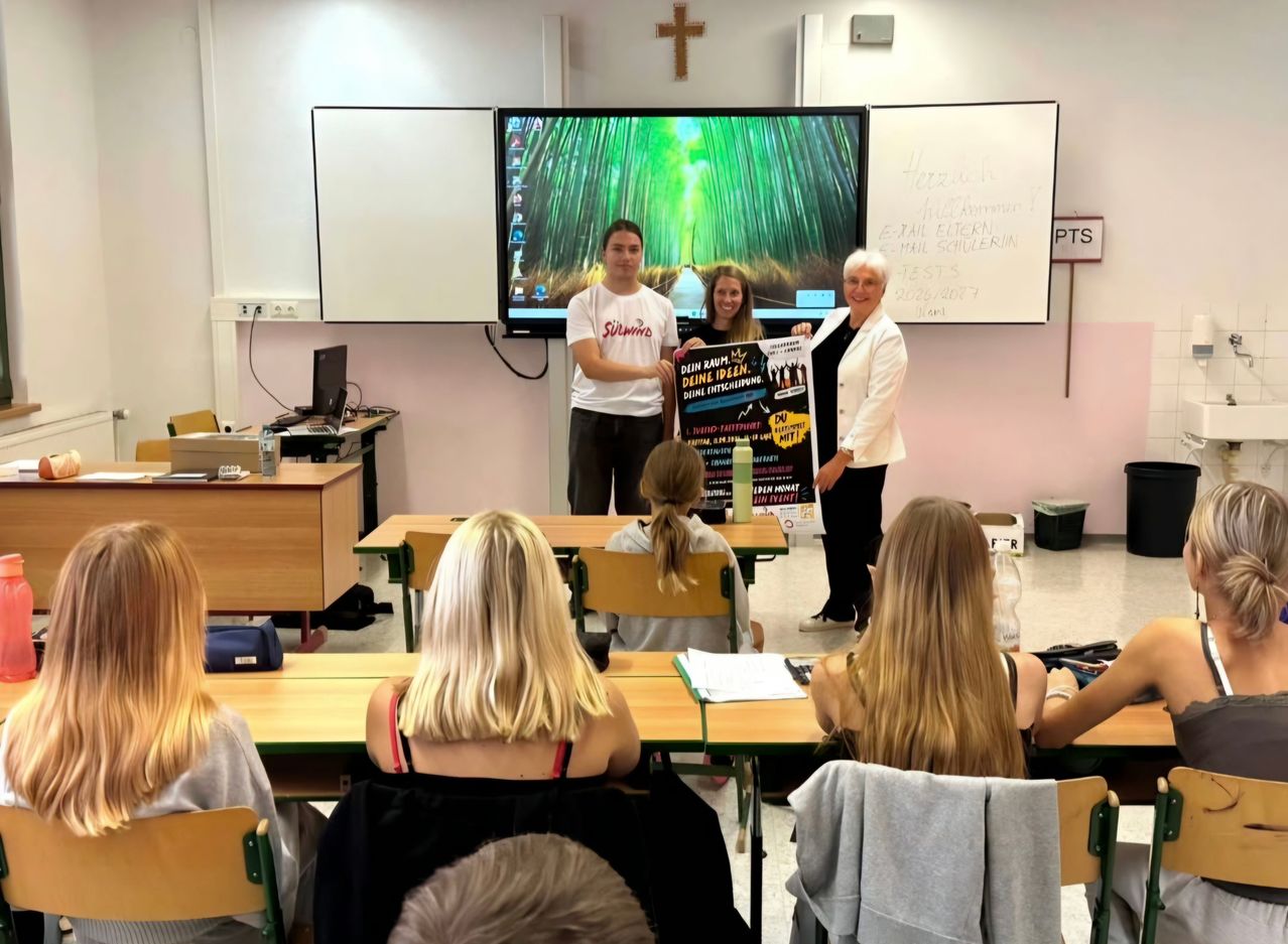 Eine Gruppe von Menschen ist in einem Klassenzimmer. Zwei Frauen und ein Mann stehen vor einer großen Leinwand. Der Mann hält ein Plakat.