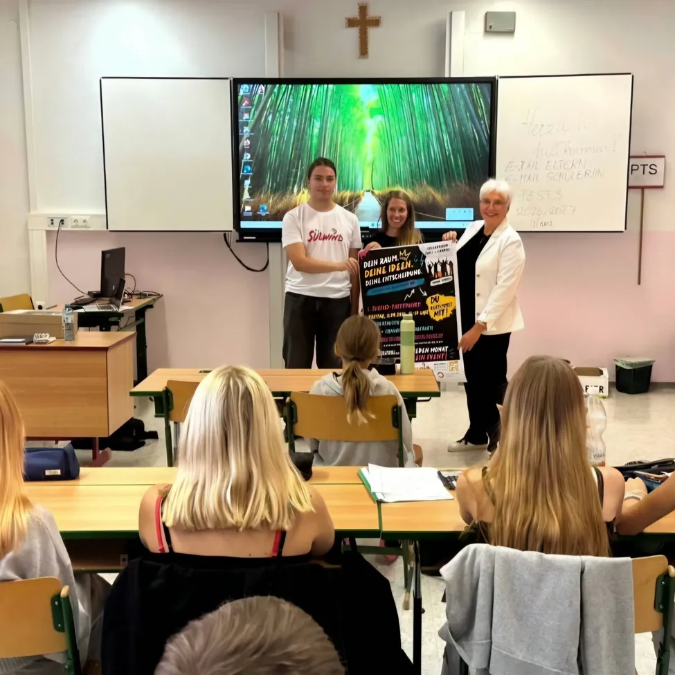 Eine Gruppe von Menschen ist in einem Klassenzimmer. Zwei Frauen und ein Mann stehen vor einer großen Leinwand. Der Mann hält ein Plakat.