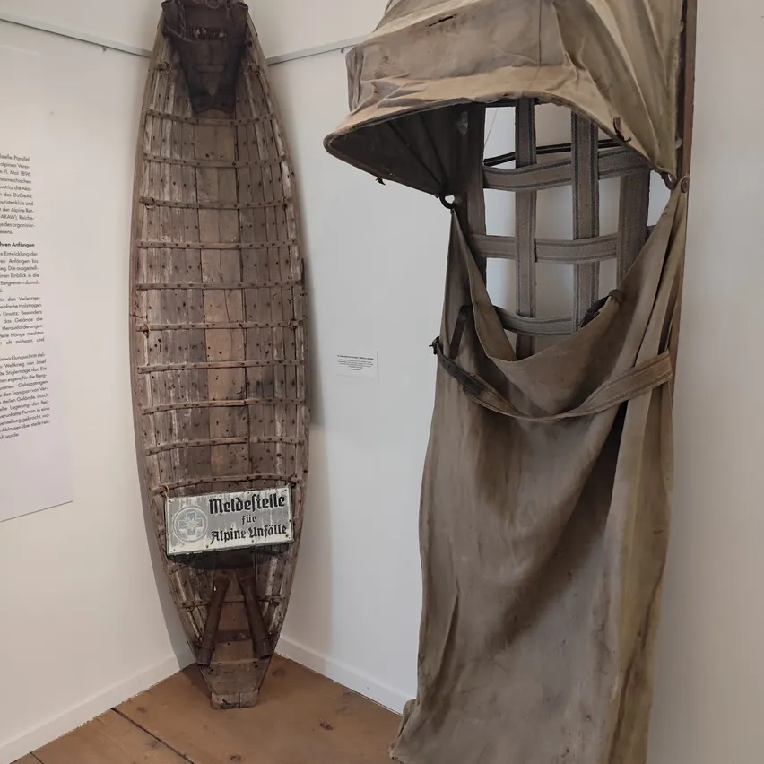 Ein altes Holzboot mit einem Metallrahmen und einem Schild, das 'Meldefelle für Alpine Unfälle' steht, ist in einem Museum auf einem Holzboden ausgestellt.