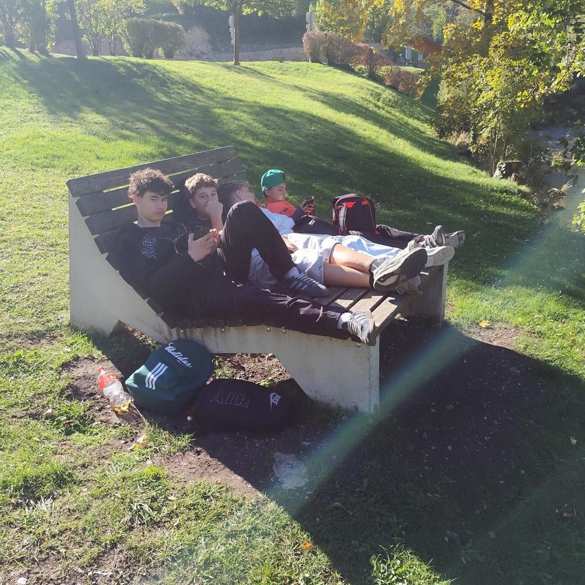 Vier junge Männer liegen auf einer Parkbank. Einer trägt einen grünen Hut und hält ein Handy.
