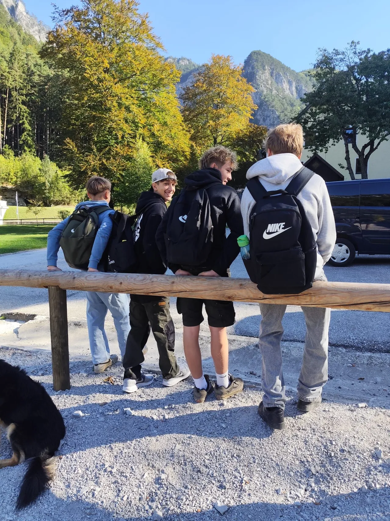 Fünf junge Männer stehen auf einem Holzzaun und schauen sich etwas an. Sie tragen alle Rucksäcke, und einer von ihnen hat eine Wasserflasche. Sie befinden sich in einem Park mit Bäumen und Bergen im Hintergrund.