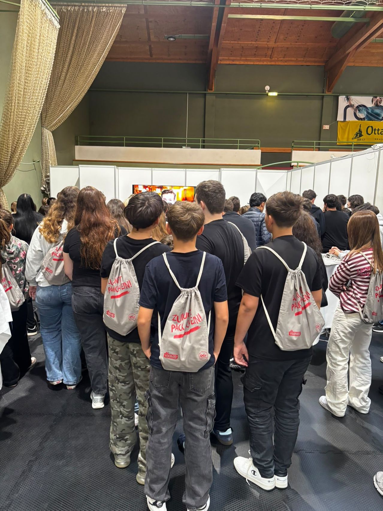 Eine Gruppe von Menschen steht in einem Raum und schaut sich eine Präsentation auf einem Bildschirm an. Der Raum ist voller Menschen, und einige von ihnen tragen Rucksäcke.