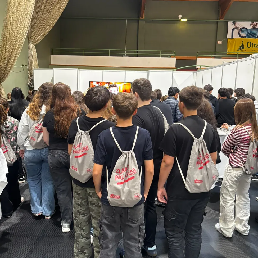 Eine Gruppe von Menschen steht in einem Raum und schaut sich eine Präsentation auf einem Bildschirm an. Der Raum ist voller Menschen, und einige von ihnen tragen Rucksäcke.