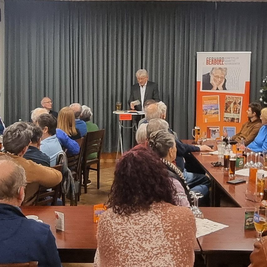 Eine Gruppe von Menschen sitzt an Tischen bei einer Versammlung, mit einem Mann am Rednerpult. Ein Schild mit dem Namen Gearbold ist sichtbar. Getränke und Papiere liegen auf den Tischen.