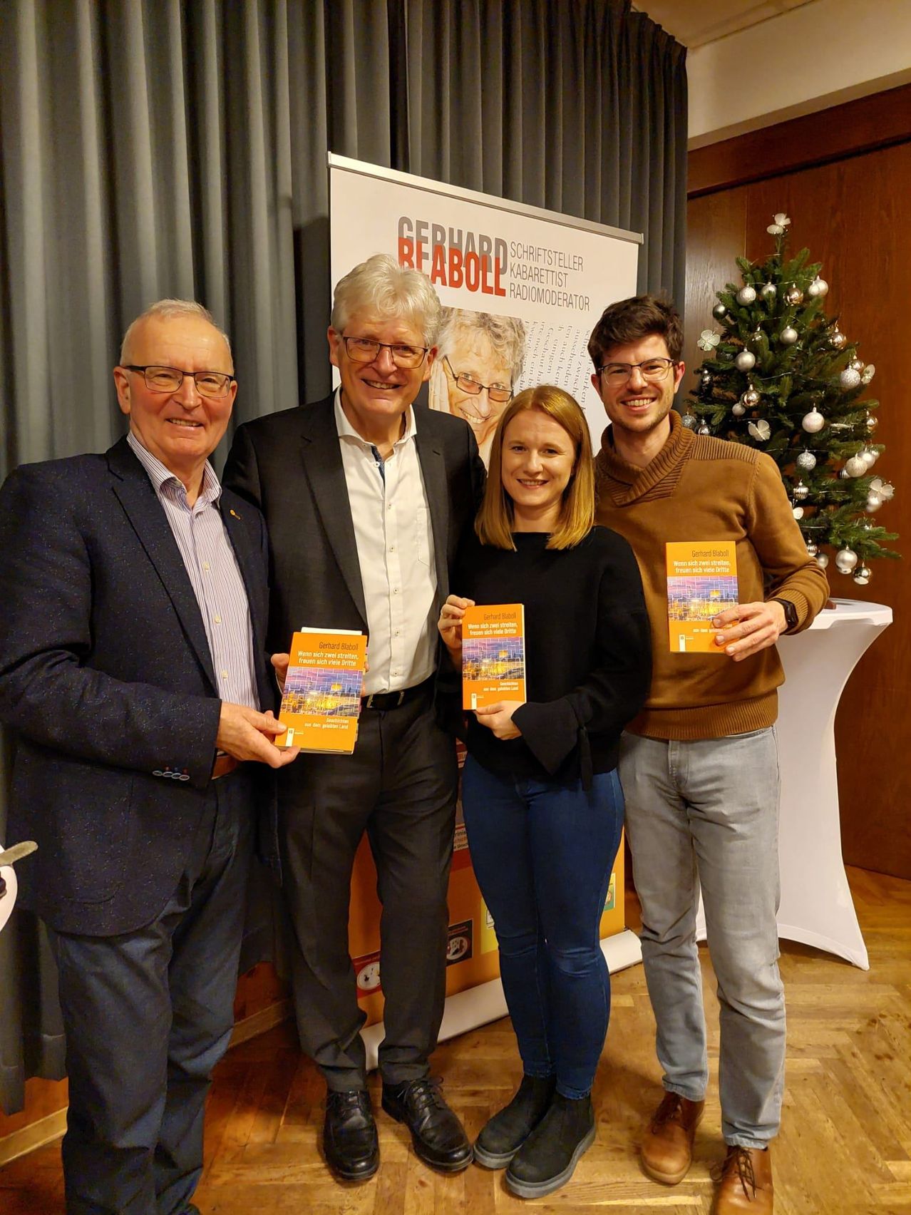 Vier Personen stehen nebeneinander und lächeln. Sie halten Bücher in den Händen. Hinter ihnen steht ein weißer Tisch und ein Weihnachtsbaum. Die Wand ist mit einem Poster dekoriert, das Text und ein Bild einer Frau enthält.
