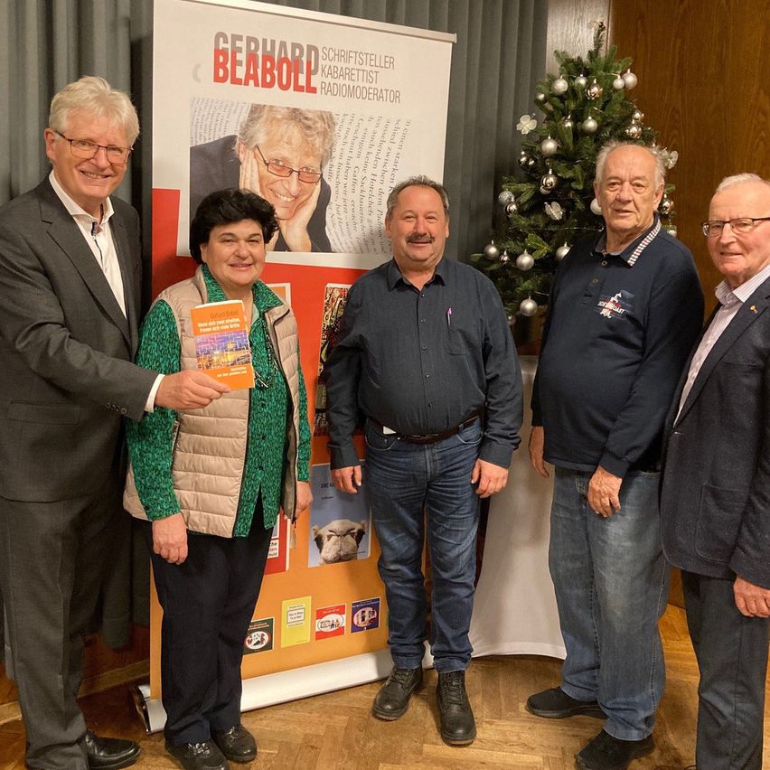 Fünf Personen stehen vor einem geschmückten Weihnachtsbaum, einer hält ein Buch. Ein Banner hinter ihnen zeigt ein Bild eines Mannes und den Text 'Gerhard Bleibold Schriftsteller Kabarettist Radiomoderator'.