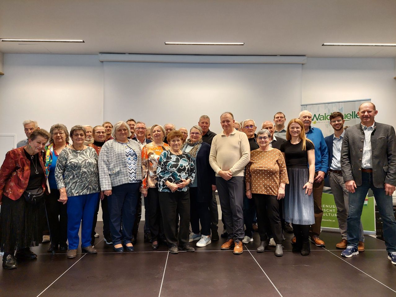 A group of people, likely a team, stands together for a photo in a conference room. The room has white walls, a green banner, and a white ceiling with lights.